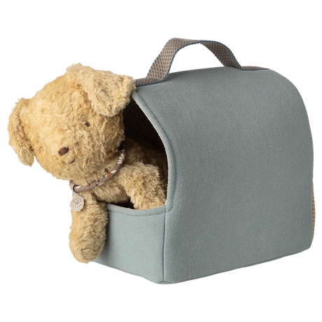 A fluffy brown teddy bear peeks out of a light blue, fabric carrier with a handle. The setting is whimsical and cute against a white background.