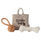 A beige fabric tote labeled “Goodie Bag” with a dog illustration is flanked by a knotted rope toy on the left and a plush bone toy on the right, set against a white background.