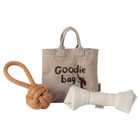 A beige fabric tote labeled “Goodie Bag” with a dog illustration is flanked by a knotted rope toy on the left and a plush bone toy on the right, set against a white background.