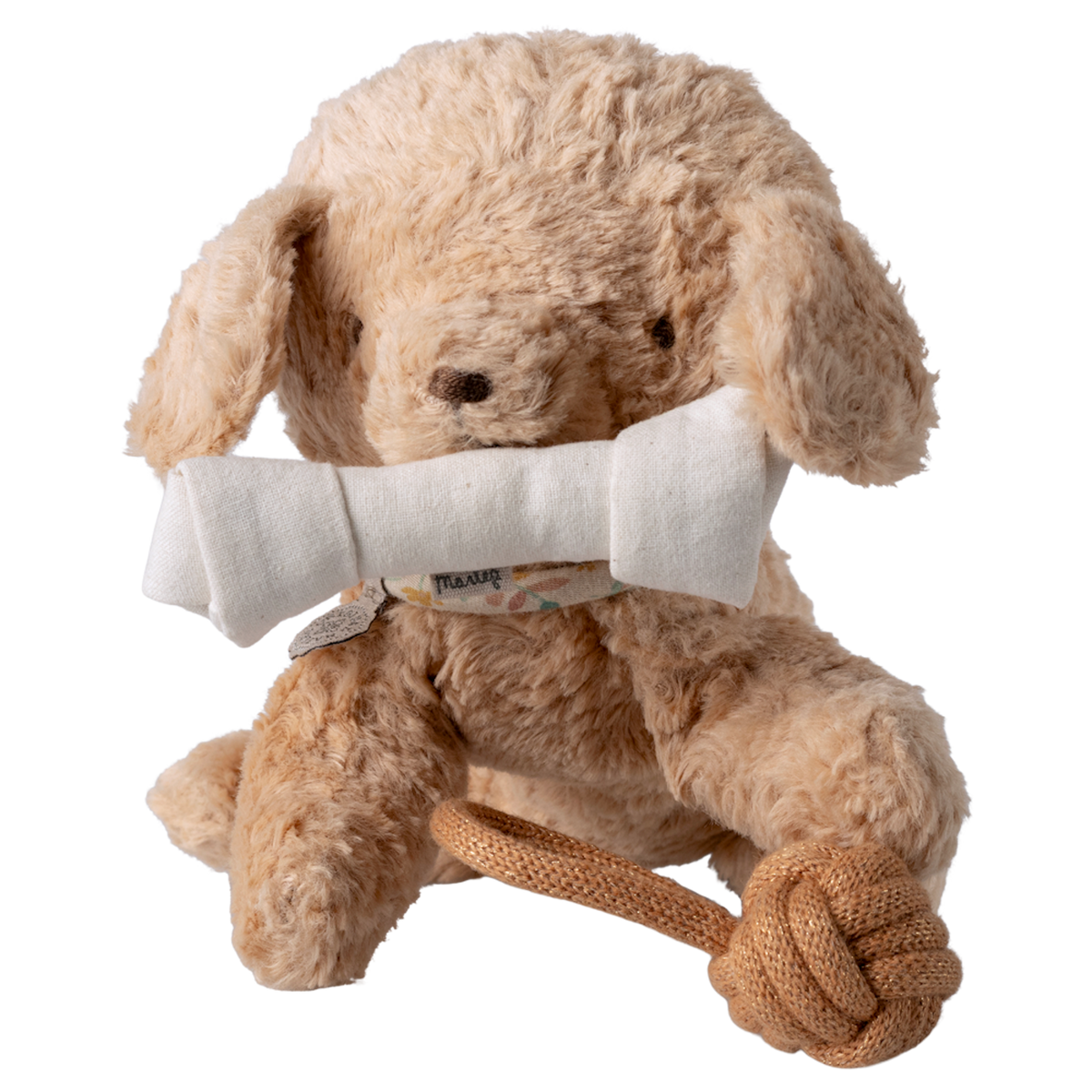 A fluffy, light brown stuffed dog sits against a white background, holding a white fabric bone in its mouth. It has a playful rope toy at its feet.