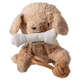 A fluffy, light brown stuffed dog sits against a white background, holding a white fabric bone in its mouth. It has a playful rope toy at its feet.