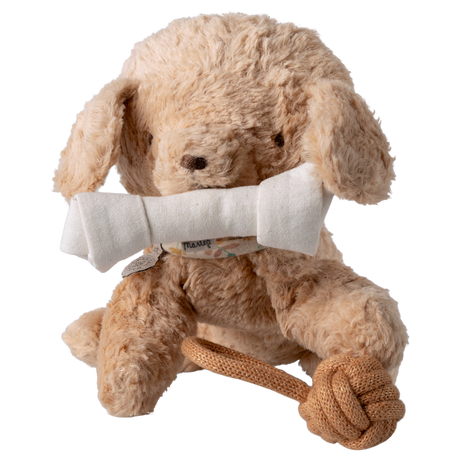 A fluffy, light brown stuffed dog sits against a white background, holding a white fabric bone in its mouth. It has a playful rope toy at its feet.