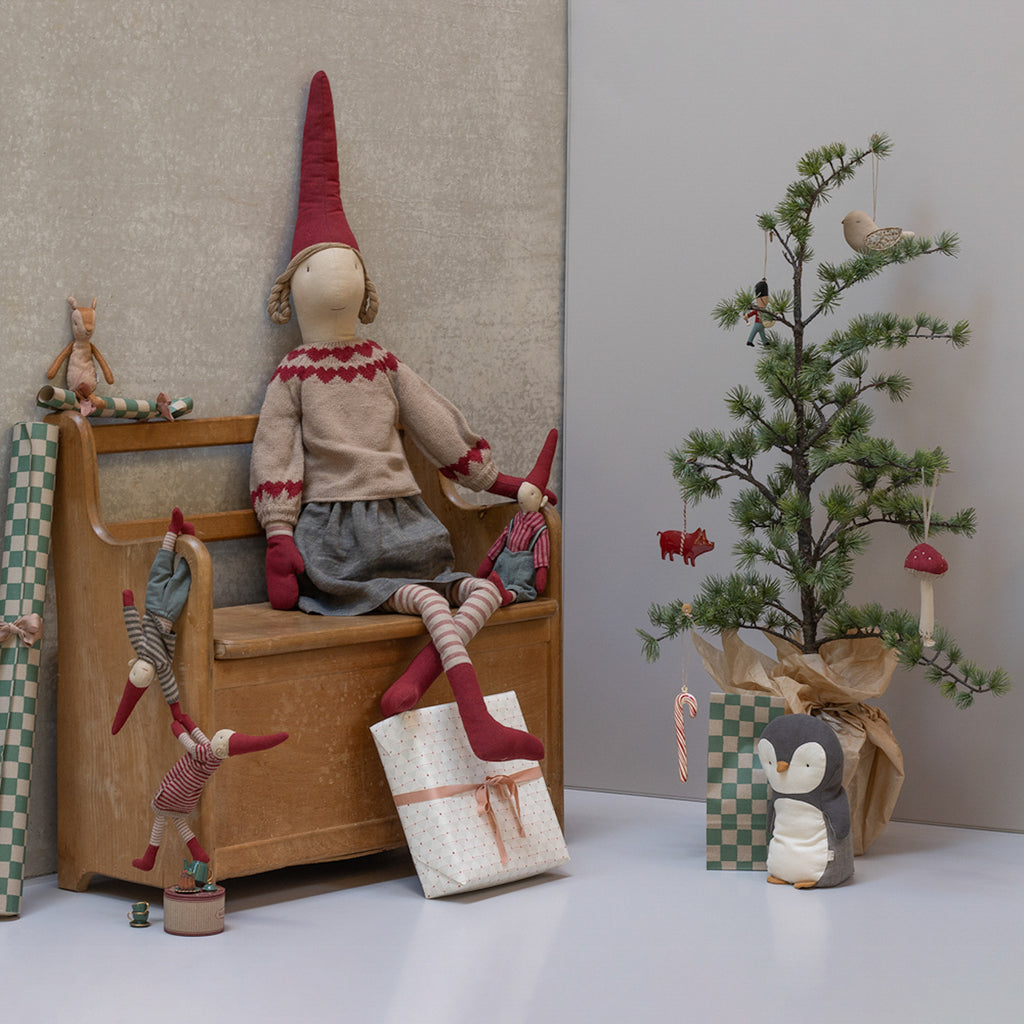 Cozy Christmas scene with soft toy elves on a wooden chair, ornaments, a small tree, and gifts. A toy penguin adds a whimsical touch.