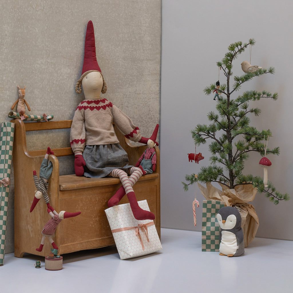 A cozy scene with a whimsical doll on a wooden bench, surrounded by playful elves. Beside them, a small decorated Christmas tree stands near wrapped gifts, evoking a festive and charming atmosphere.