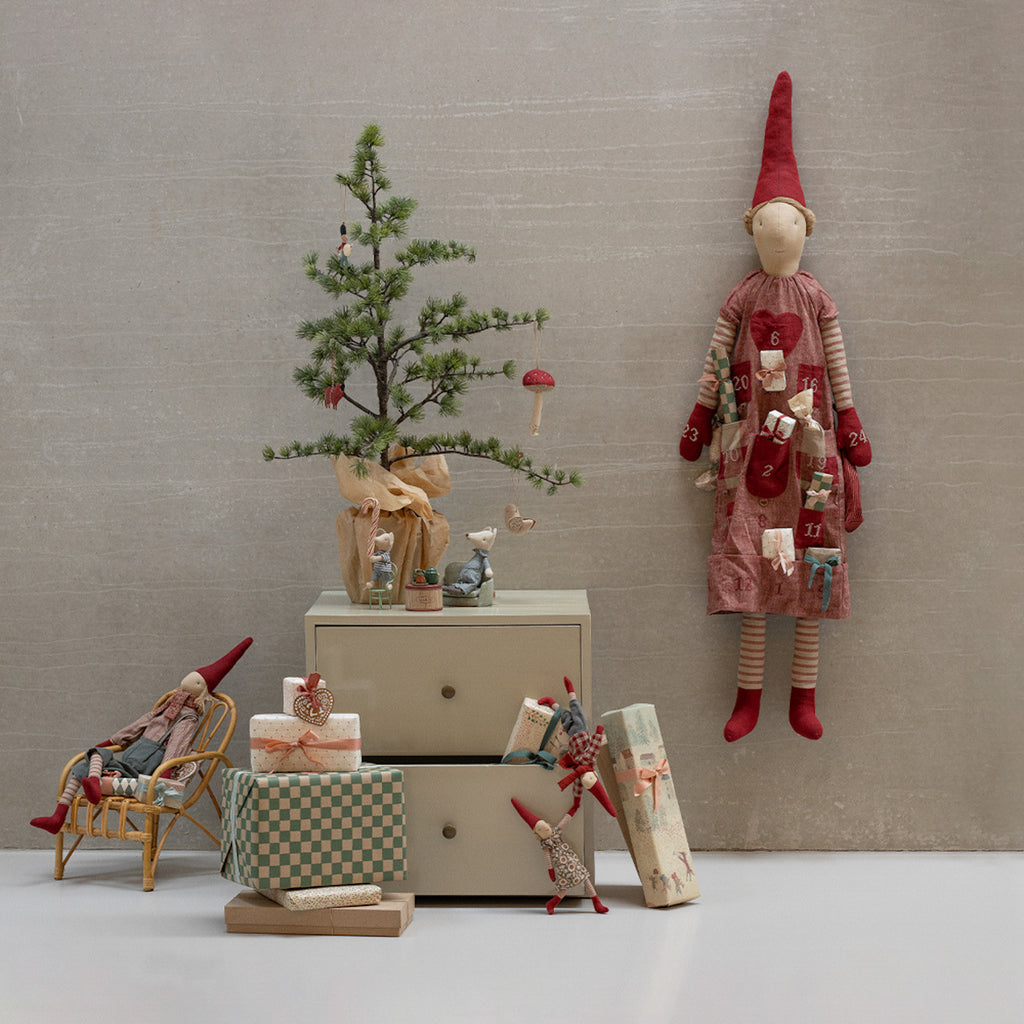 A festive scene with a small, decorated pine tree on a cabinet. An Advent calendar doll with red hat and mittens hangs nearby. Wrapped gifts, a small seated figure, and playful mice add whimsical touches.
