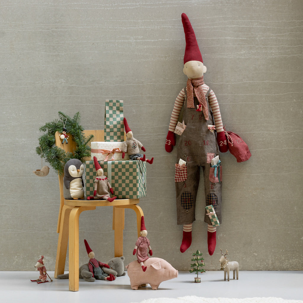 A whimsical holiday scene with a stuffed gnome wearing overalls and a red hat hanging on a wall. Below are festive toys, a chair with gifts, and a small tree.