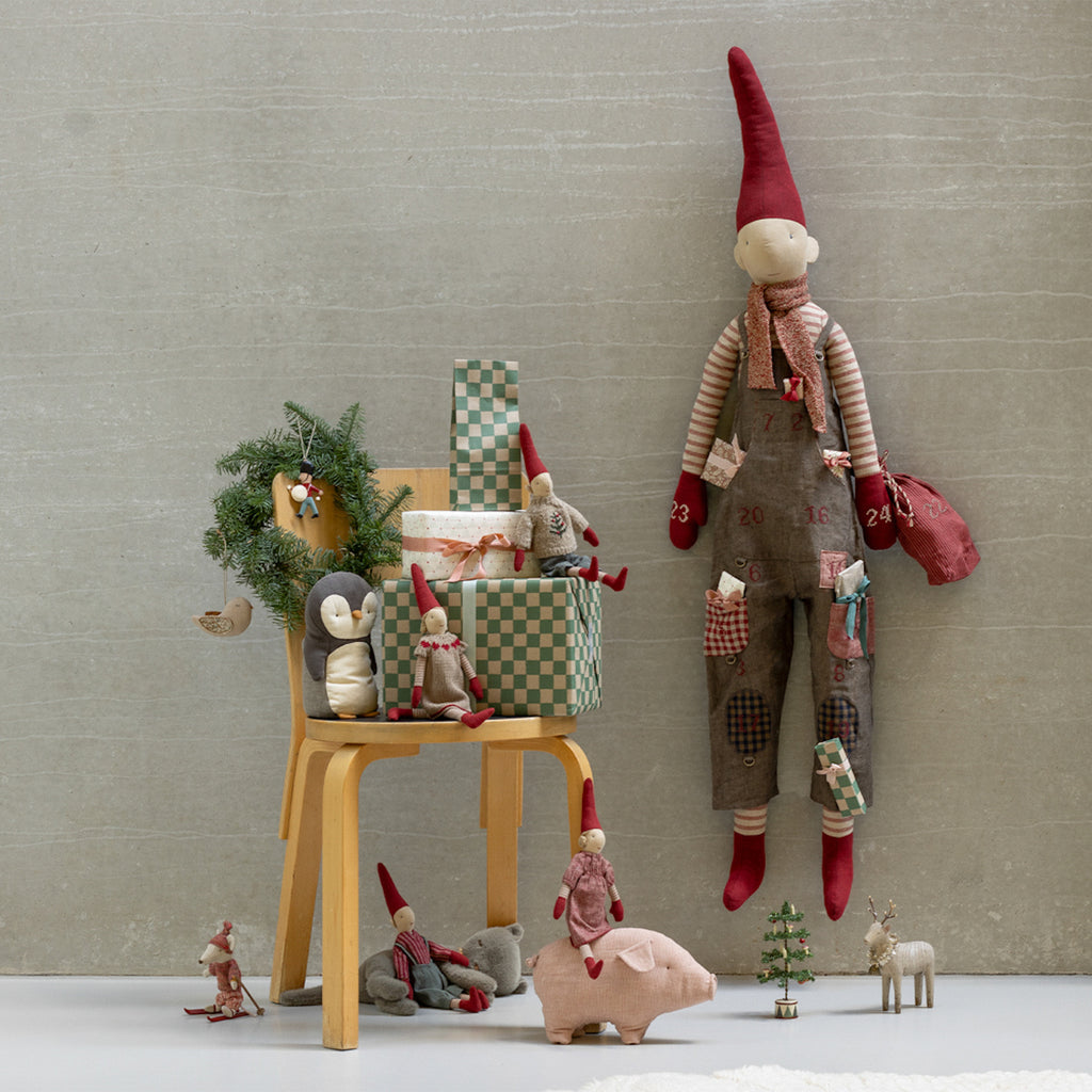 Festive scene with a tall, whimsical elf doll in overalls beside a chair holding wrapped gifts and small plush toys. A cozy, holiday mood is conveyed.