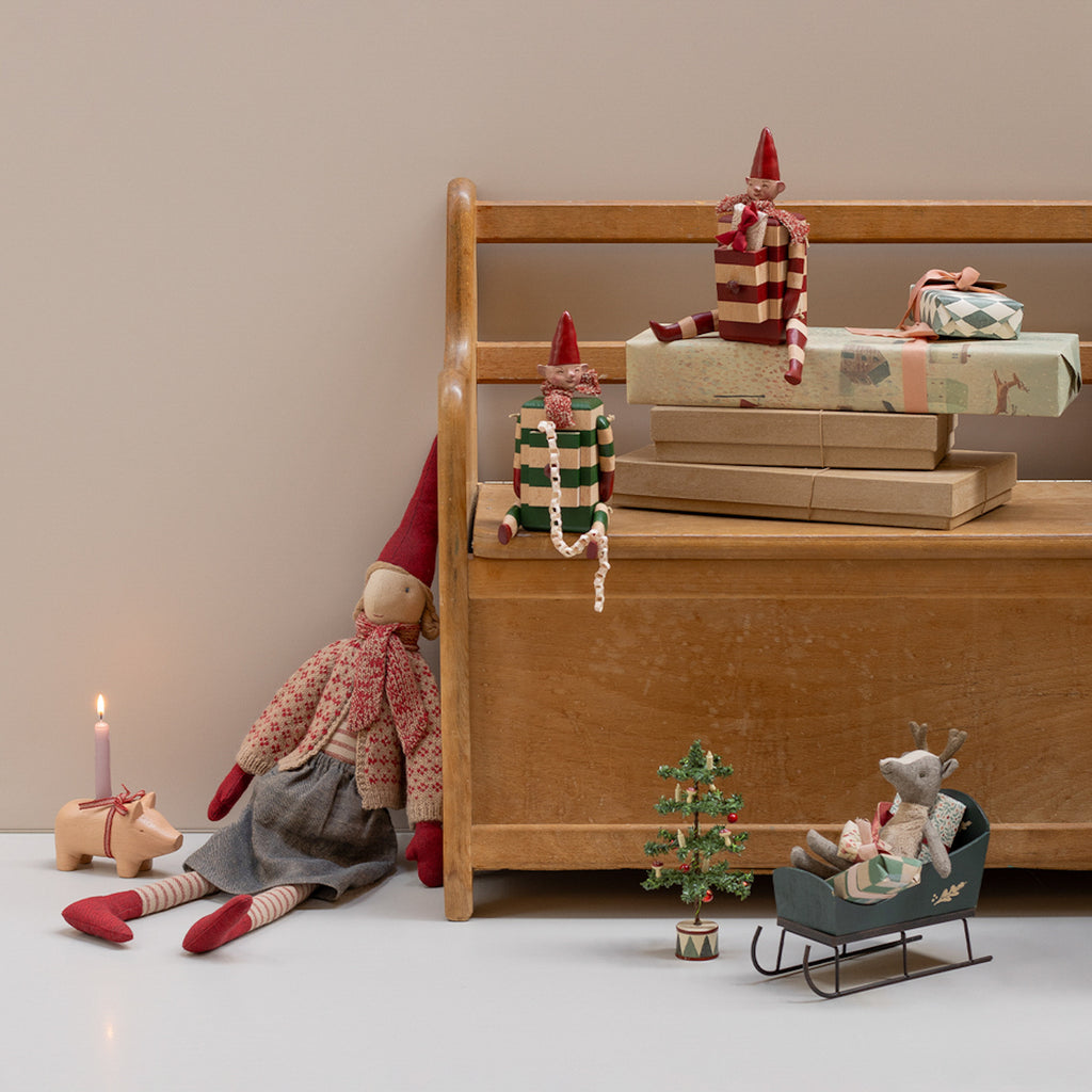 Whimsical Christmas scene with small elf dolls on a wooden bench, wrapped gifts, a toy reindeer in a sleigh, and a mini Christmas tree. Cozy and festive.