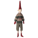 Toy doll in a dark setting, with a tall red hat, striped sweater featuring "24," gray pants, red gloves, striped socks, and red shoes, conveying whimsy.
