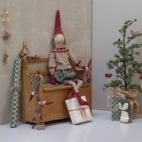 A festive scene with plush dolls in red hats seated on a wooden bench. A small decorated tree stands beside a wrapped gift and a penguin plush. Cozy and whimsical.