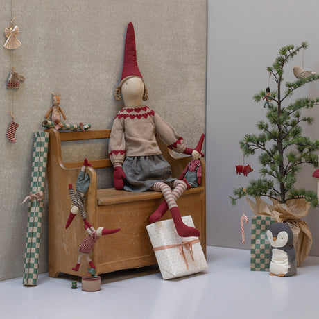 A festive scene with plush dolls in red hats seated on a wooden bench. A small decorated tree stands beside a wrapped gift and a penguin plush. Cozy and whimsical.