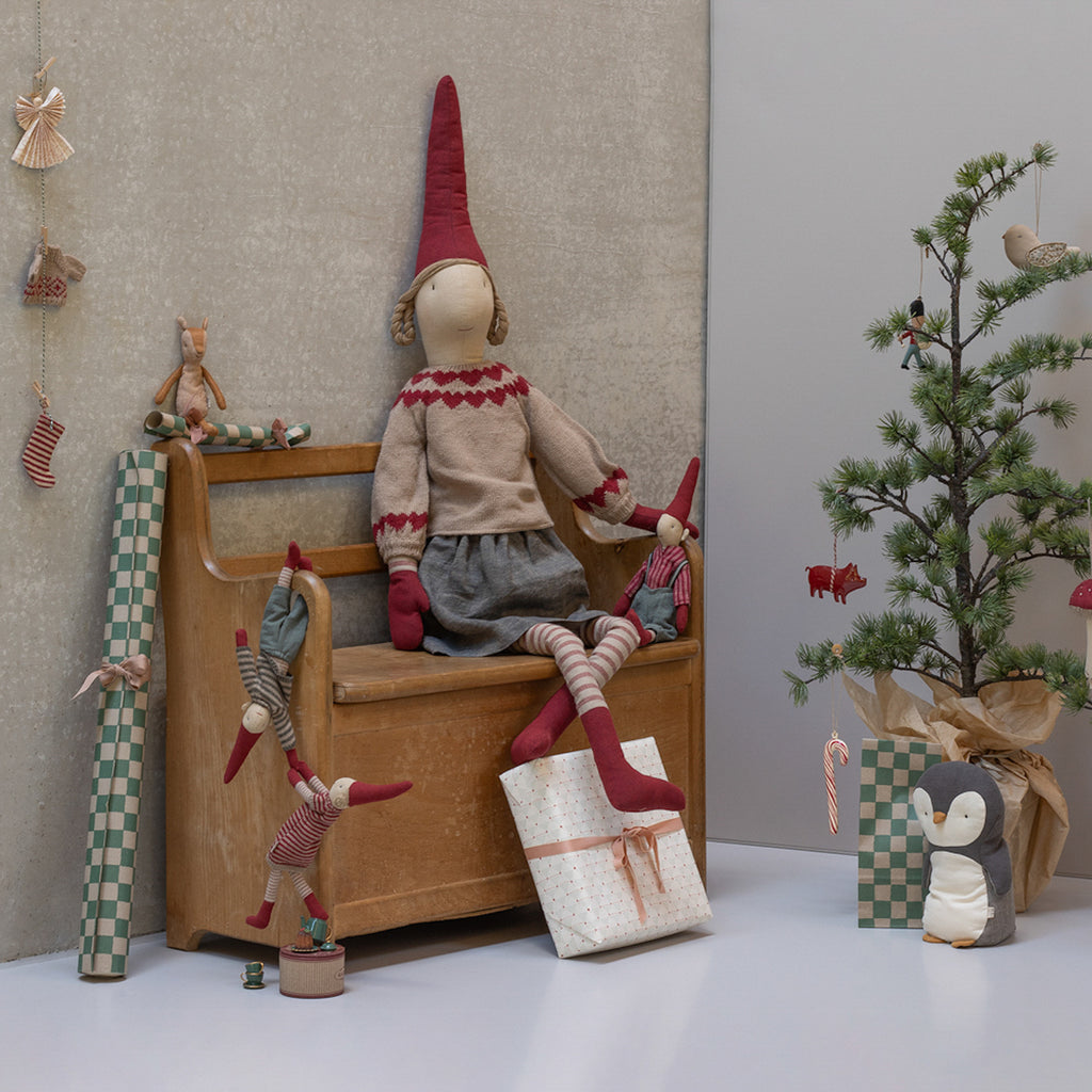 A festive scene with plush dolls in red hats seated on a wooden bench. A small decorated tree stands beside a wrapped gift and a penguin plush. Cozy and whimsical.