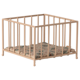A small, beige metal playpen with a colorful, quilted cushion sits empty against a white background, conveying a sense of simplicity and nostalgia.