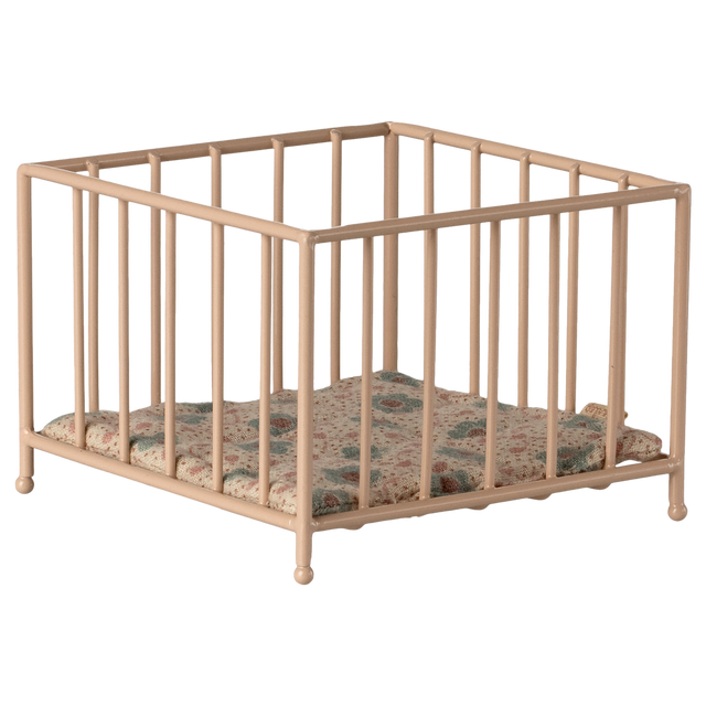 A small, beige metal playpen with a colorful, quilted cushion sits empty against a white background, conveying a sense of simplicity and nostalgia.