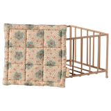 Vintage-style beige and blue floral quilted pillow leaning against a small, light brown wooden crib on a white background. The scene feels nostalgic and cozy.