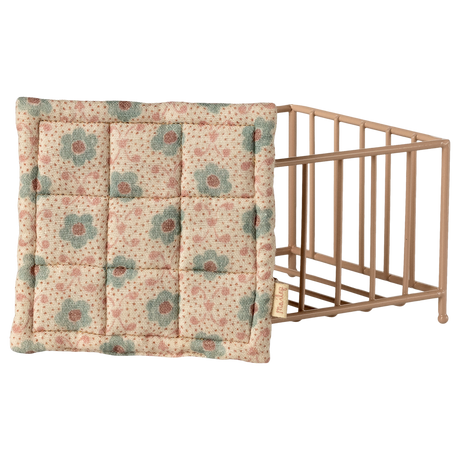 Vintage-style beige and blue floral quilted pillow leaning against a small, light brown wooden crib on a white background. The scene feels nostalgic and cozy.