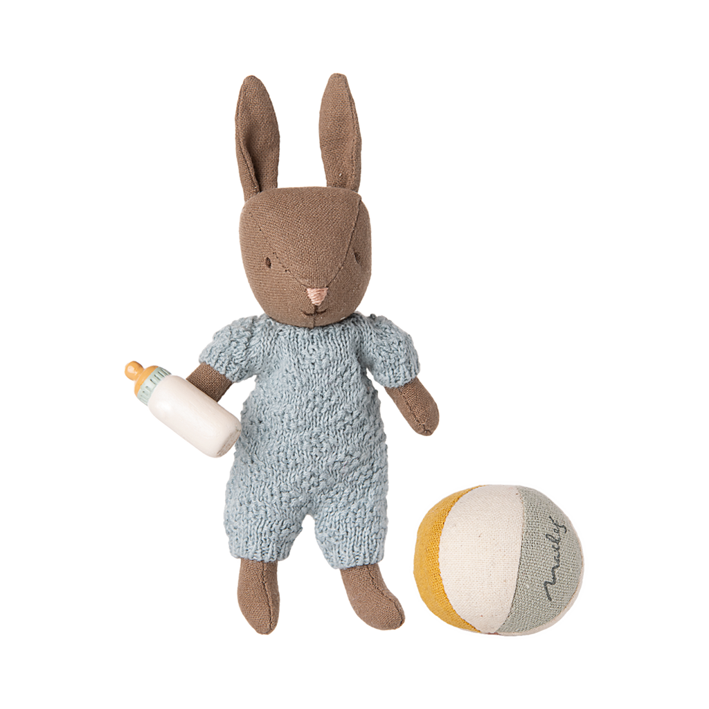A cute brown fabric bunny in a blue knitted outfit holds a white baby bottle, next to a colorful patchwork ball, on a plain background.
