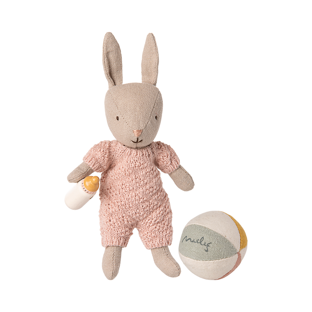 A cute fabric bunny wearing a pink knitted outfit holds a small milk bottle. A soft, multicolored ball inscribed with "Maileg" is nearby. Playful and cozy vibe.