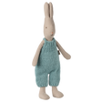 A beige stuffed bunny stands upright, wearing blue knitted overalls. Its simple, minimalistic design conveys a cozy, charming feel against a white background.