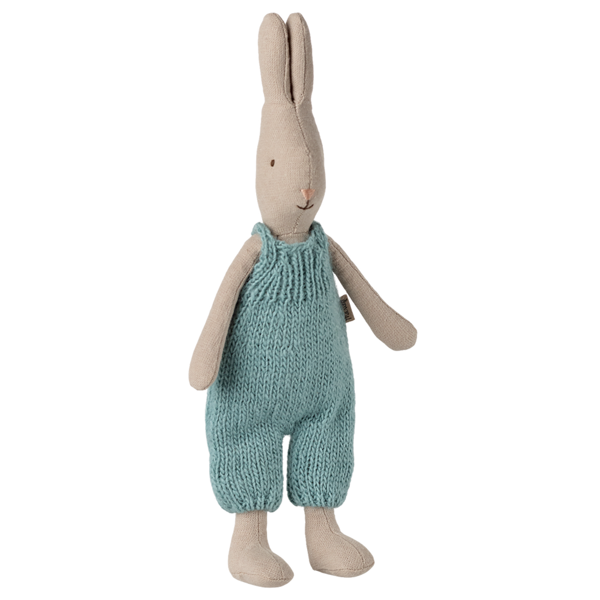 A beige stuffed bunny stands upright, wearing blue knitted overalls. Its simple, minimalistic design conveys a cozy, charming feel against a white background.