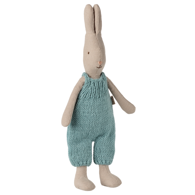 A beige stuffed bunny stands upright, wearing blue knitted overalls. Its simple, minimalistic design conveys a cozy, charming feel against a white background.