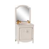 A miniature white vanity with a mirror, featuring a gold faucet and taps. The cabinet door is slightly ajar, conveying a quaint and elegant feel.