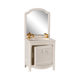 A miniature white vanity with a mirror, featuring a gold faucet and taps. The cabinet door is slightly ajar, conveying a quaint and elegant feel.