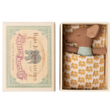 A small brown stuffed mouse rests in an open box, nestled in a yellow-patterned blanket. The box lid features an illustration of an elephant and reads “Baby Jumbo Matches.”