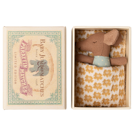 A small brown stuffed mouse rests in an open box, nestled in a yellow-patterned blanket. The box lid features an illustration of an elephant and reads “Baby Jumbo Matches.”