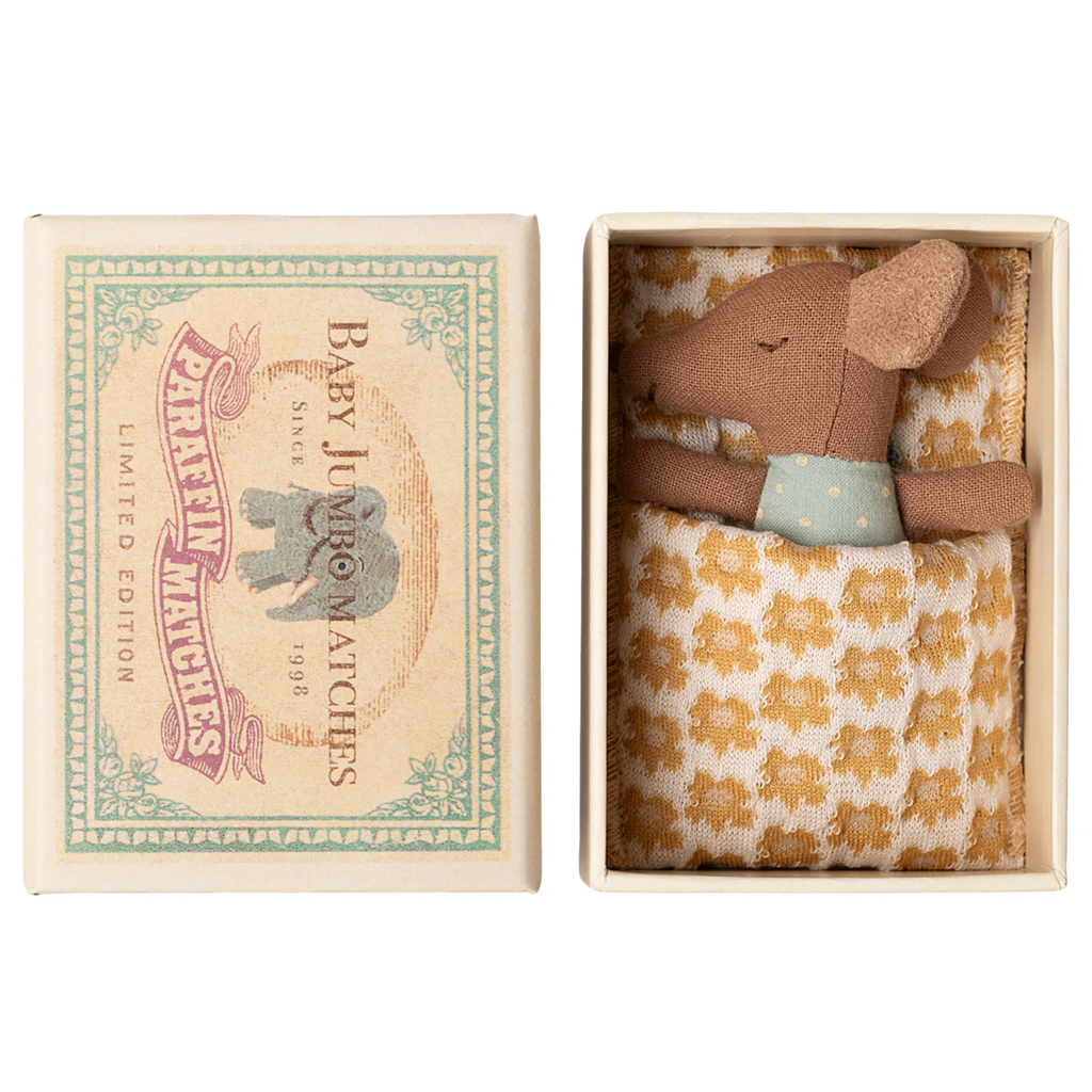 A small brown stuffed mouse rests in an open box, nestled in a yellow-patterned blanket. The box lid features an illustration of an elephant and reads “Baby Jumbo Matches.”