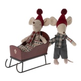 Two whimsical toy mice in red hats and cozy scarves are on a sled. One sits in the sled, the other stands beside, both appearing joyful and festive.