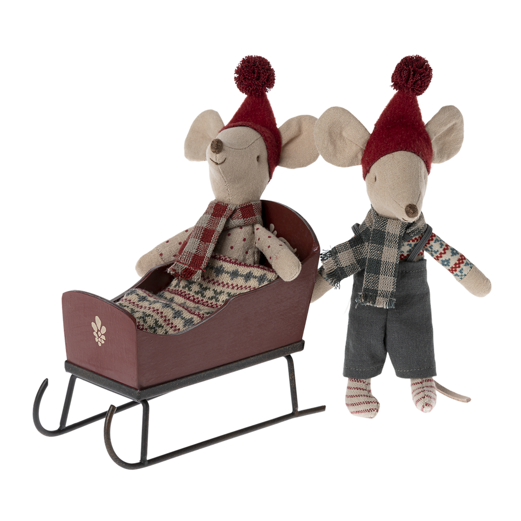 Two whimsical toy mice in red hats and cozy scarves are on a sled. One sits in the sled, the other stands beside, both appearing joyful and festive.