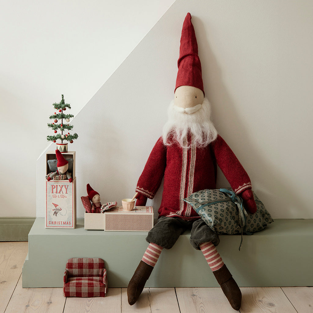 Large Santa doll in a red coat and hat sits smiling, surrounded by mini Santa figures in boxes, a small Christmas tree, and a plaid cushion. Cozy holiday scene.
