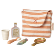 A small striped fabric pouch with a red cross is surrounded by a collection of miniature toiletries: a white cup, a perfume bottle, a toothbrush, a toothpaste tube, and a golden hand mirror, all against a white  background.
