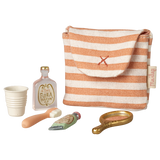 A small striped fabric pouch with a red cross is surrounded by a collection of miniature toiletries: a white cup, a perfume bottle, a toothbrush, a toothpaste tube, and a golden hand mirror, all against a white  background.