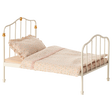 Antique-style miniature white metal bed with curved headboard and footboard, adorned with golden accents. Covered in polka-dot bedding and ruffled pillow.