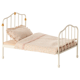 Antique-style miniature white metal bed with curved headboard and footboard, adorned with golden accents. Covered in polka-dot bedding and ruffled pillow.