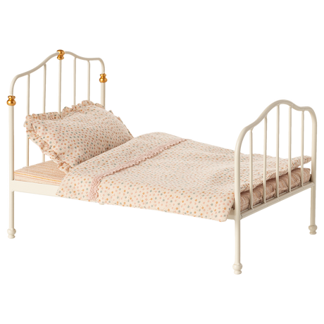 Antique-style miniature white metal bed with curved headboard and footboard, adorned with golden accents. Covered in polka-dot bedding and ruffled pillow.