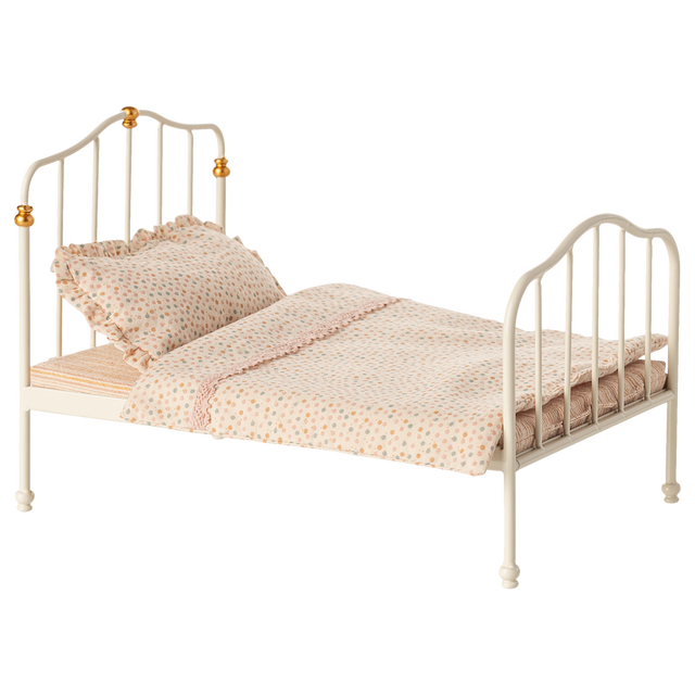 Antique-style miniature white metal bed with curved headboard and footboard, adorned with golden accents. Covered in polka-dot bedding and ruffled pillow.