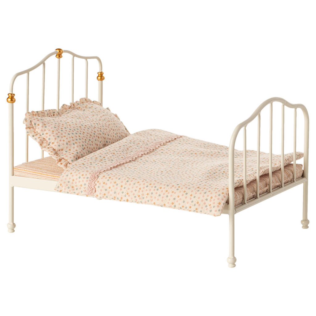 Antique-style miniature white metal bed with curved headboard and footboard, adorned with golden accents. Covered in polka-dot bedding and ruffled pillow.