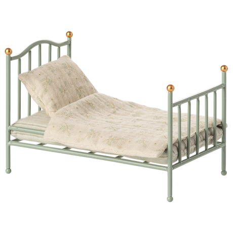 A miniature, vintage-style metal bed with a pale green frame and golden accents. It has a cream-colored, patterned quilt and pillow, conveying a nostalgic charm.