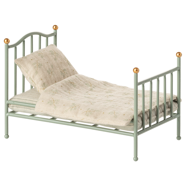 A miniature, vintage-style metal bed with a pale green frame and golden accents. It has a cream-colored, patterned quilt and pillow, conveying a nostalgic charm.