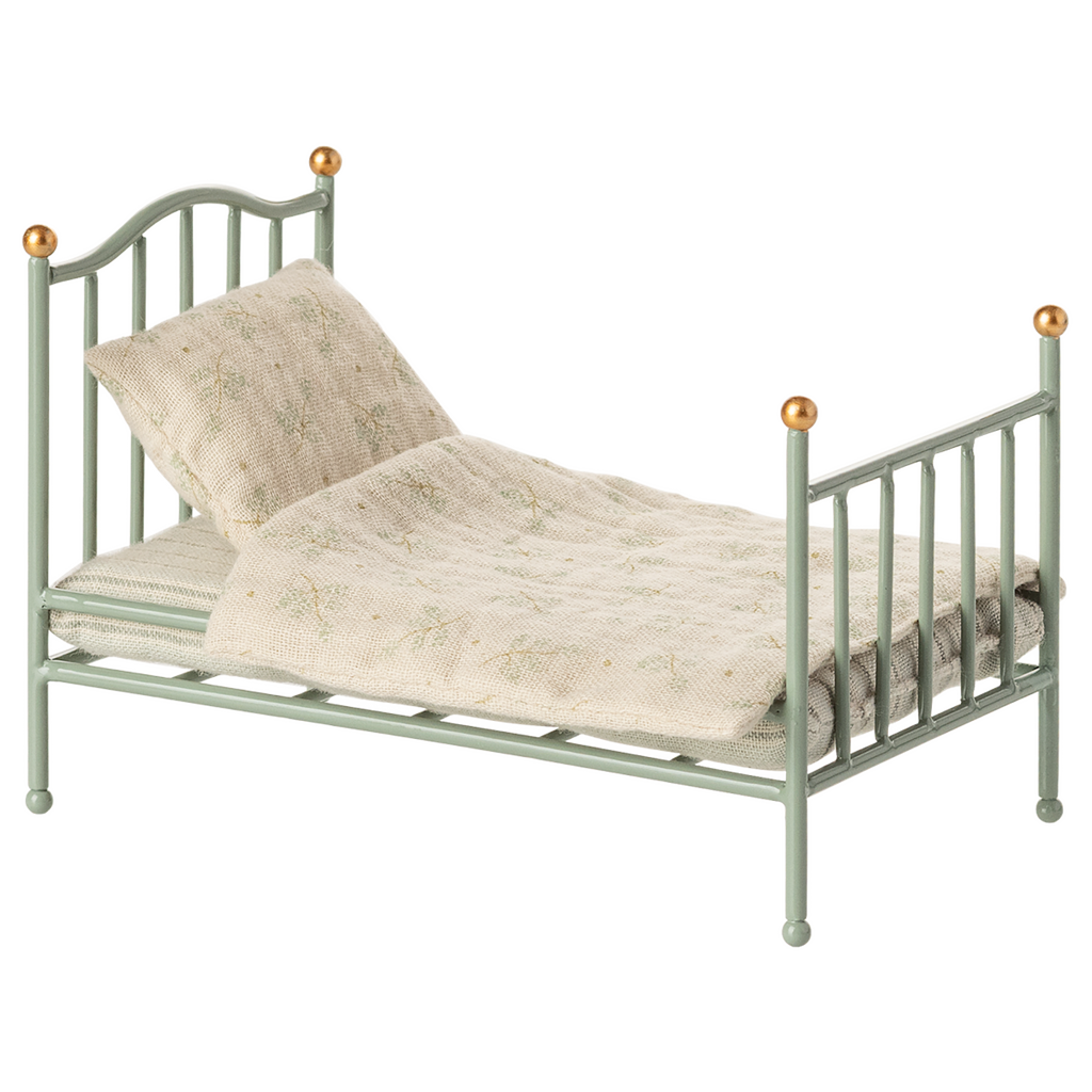 A miniature, vintage-style metal bed with a pale green frame and golden accents. It has a cream-colored, patterned quilt and pillow, conveying a nostalgic charm.