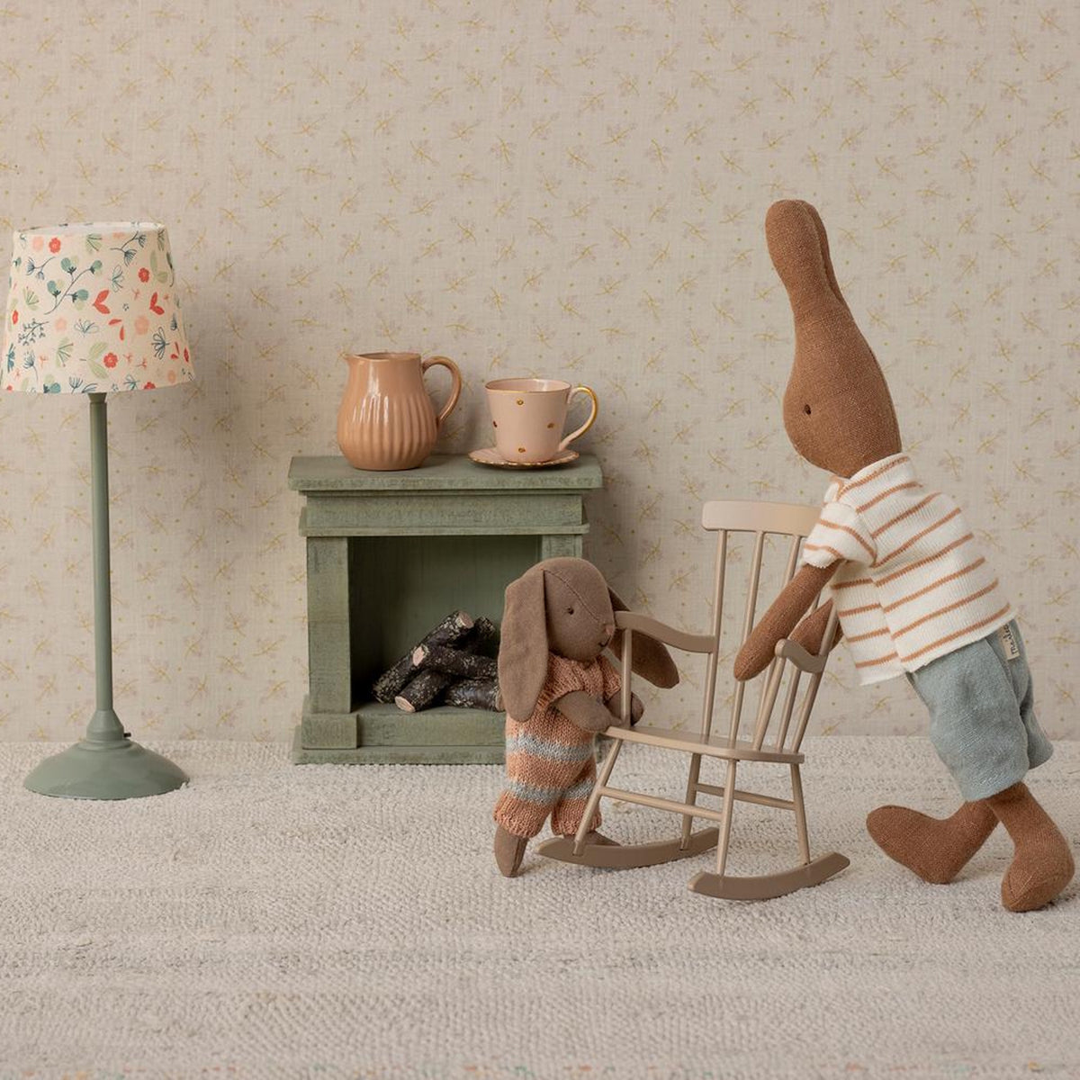 Two Maileg brown bunny dolls in a miniature room with a rocking chair, fireplace, and floral lamp.