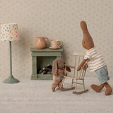 Two Maileg brown bunny dolls in a miniature room with a rocking chair, fireplace, and floral lamp.