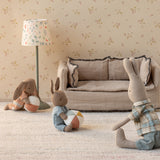 Three Maileg bunny dolls playing with balls in a miniature living room with a linen sofa and floral lamp.