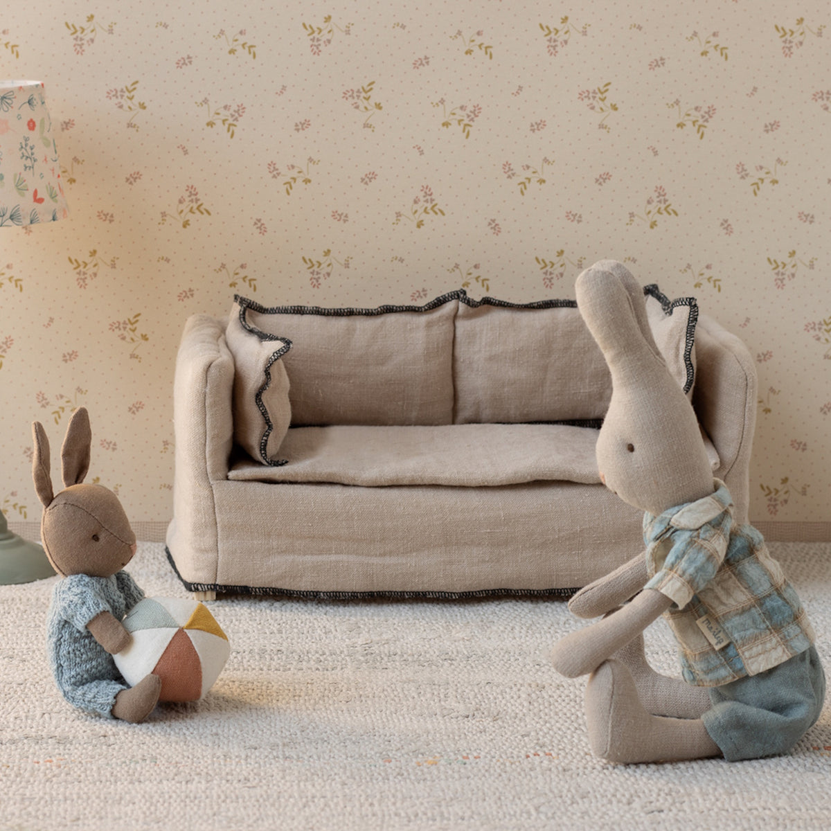 Two Maileg rabbit plush toys playing with a ball in a miniature living room with a linen sofa and floral wallpaper.