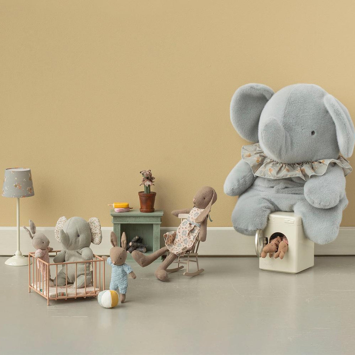 Maileg plush rabbits and elephants in a miniature playroom with a toy crib, rocking chair, and washing machine.