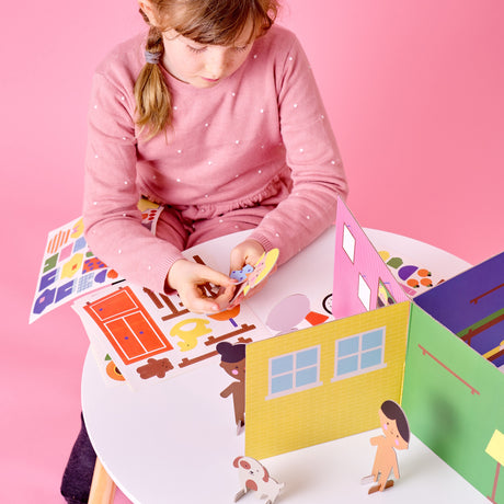 Young girl in a pink sweater applying stickers to character figures at a table with a 3D paper house playset.