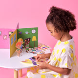 Young girl with curly hair peeling stickers to decorate paper characters and a 3D house playset.